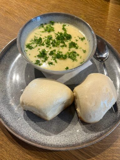 Eine Schüssel cremige Kartoffelsuppe mit Kräutern und zwei Dampfnudeln auf einem Teller.