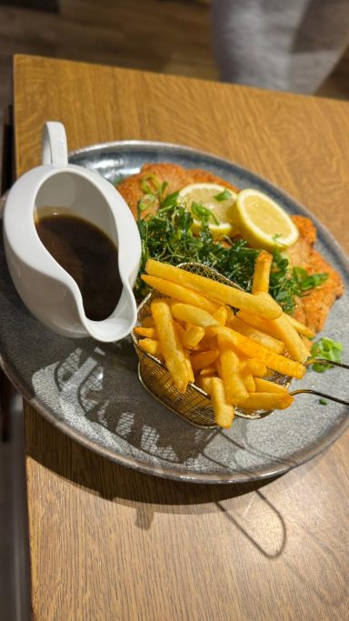 Ein Teller mit paniertem Schnitzel, Pommes und Zitronenscheiben, dazu eine Sauce in einem Kännchen.